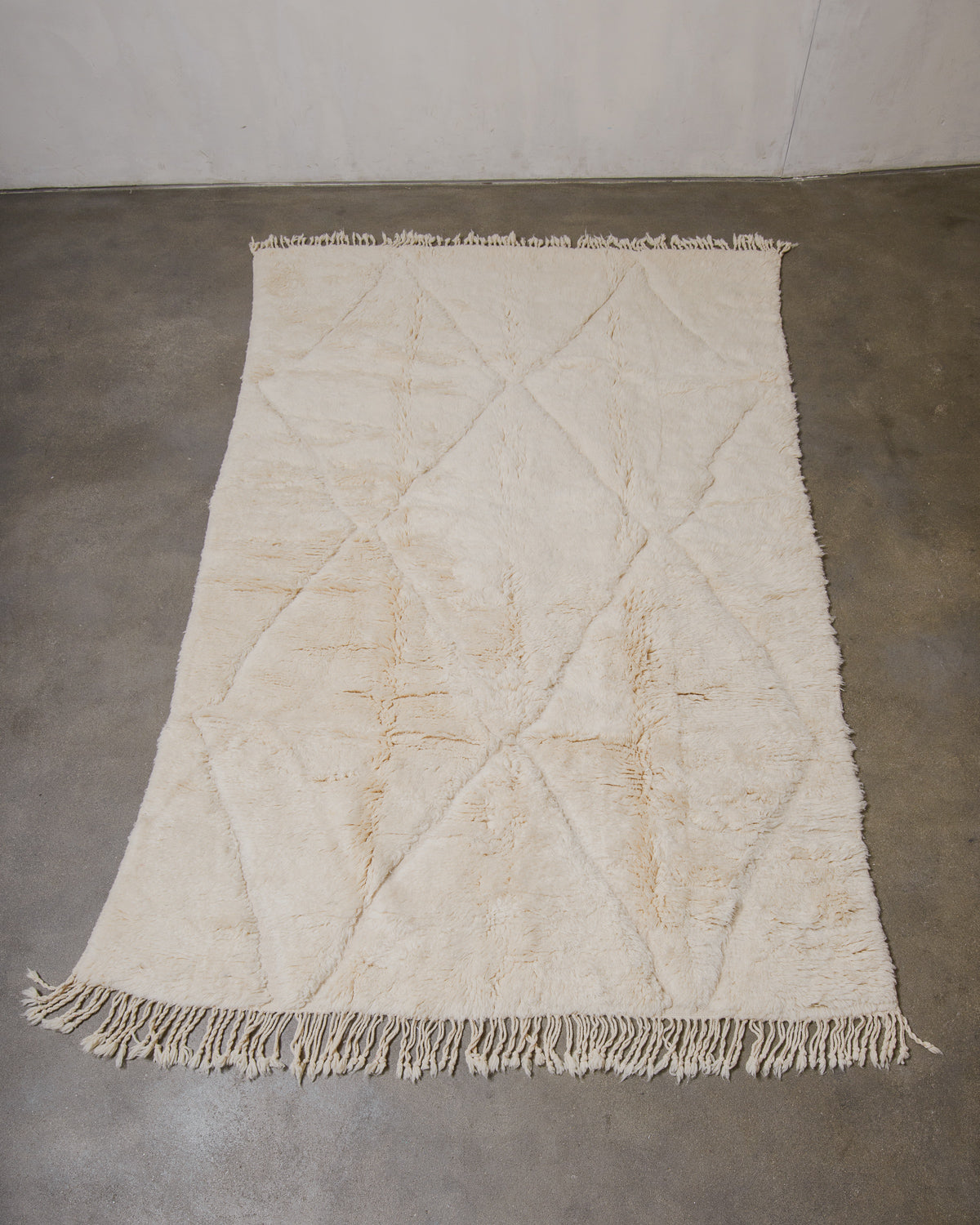 Beige woven wool rug with diamond pattern and fringe on a concrete floor.